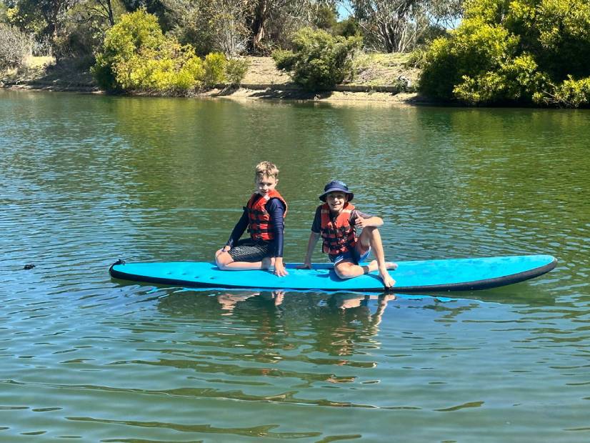 Two kids sitting on surf board on water at Lake Dewar Y Discovery camp